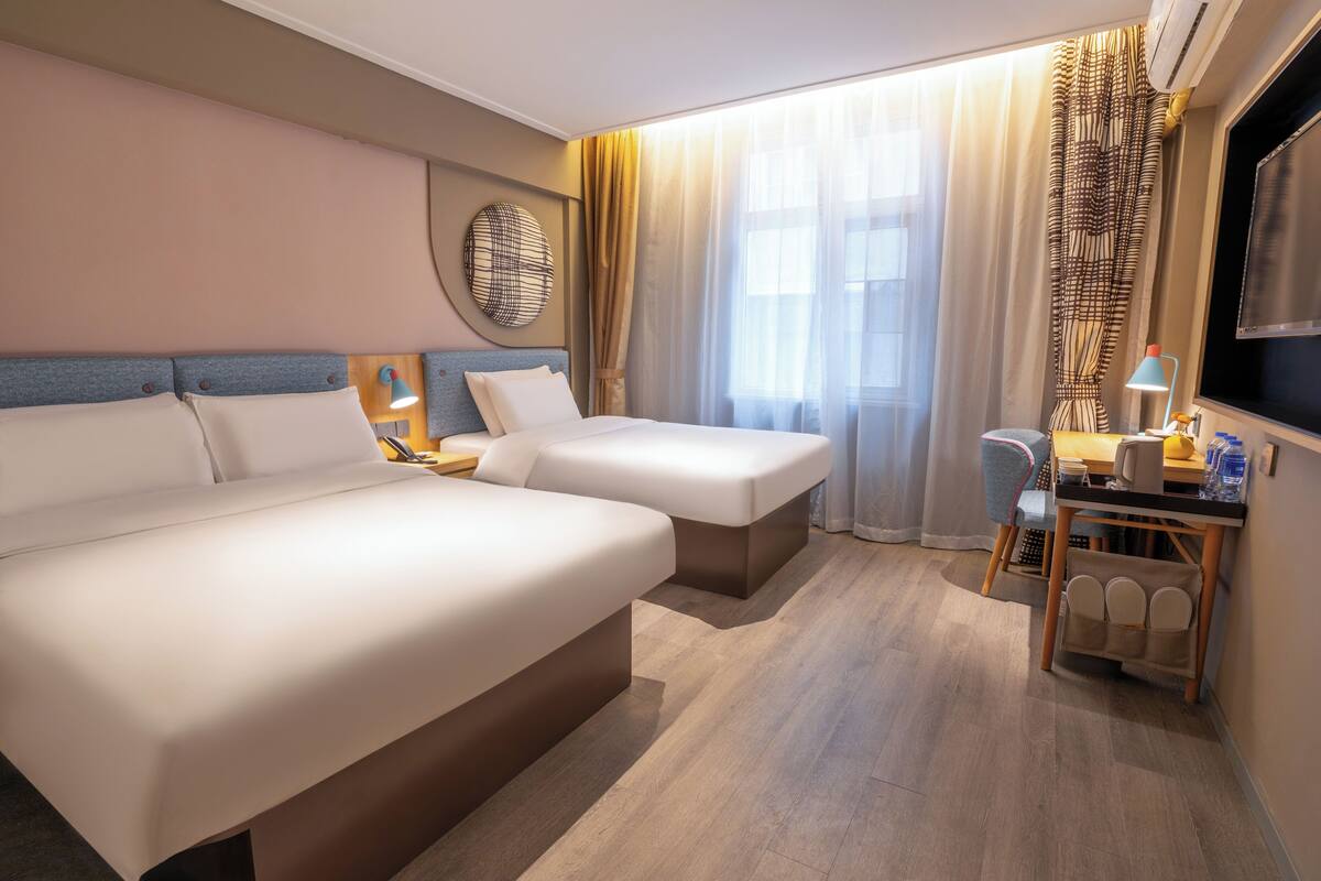 A hotel room with two beds, a desk, a chair, a TV, and a large window with curtains.