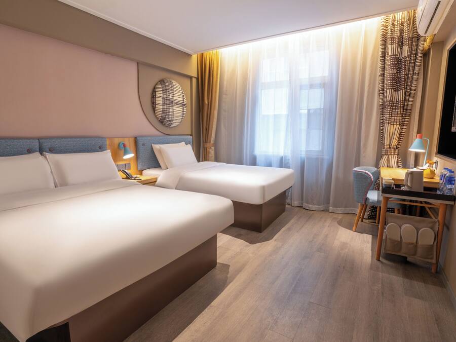 Habitación de hotel con dos camas, un escritorio con silla, televisor y un ventanal con cortinas.