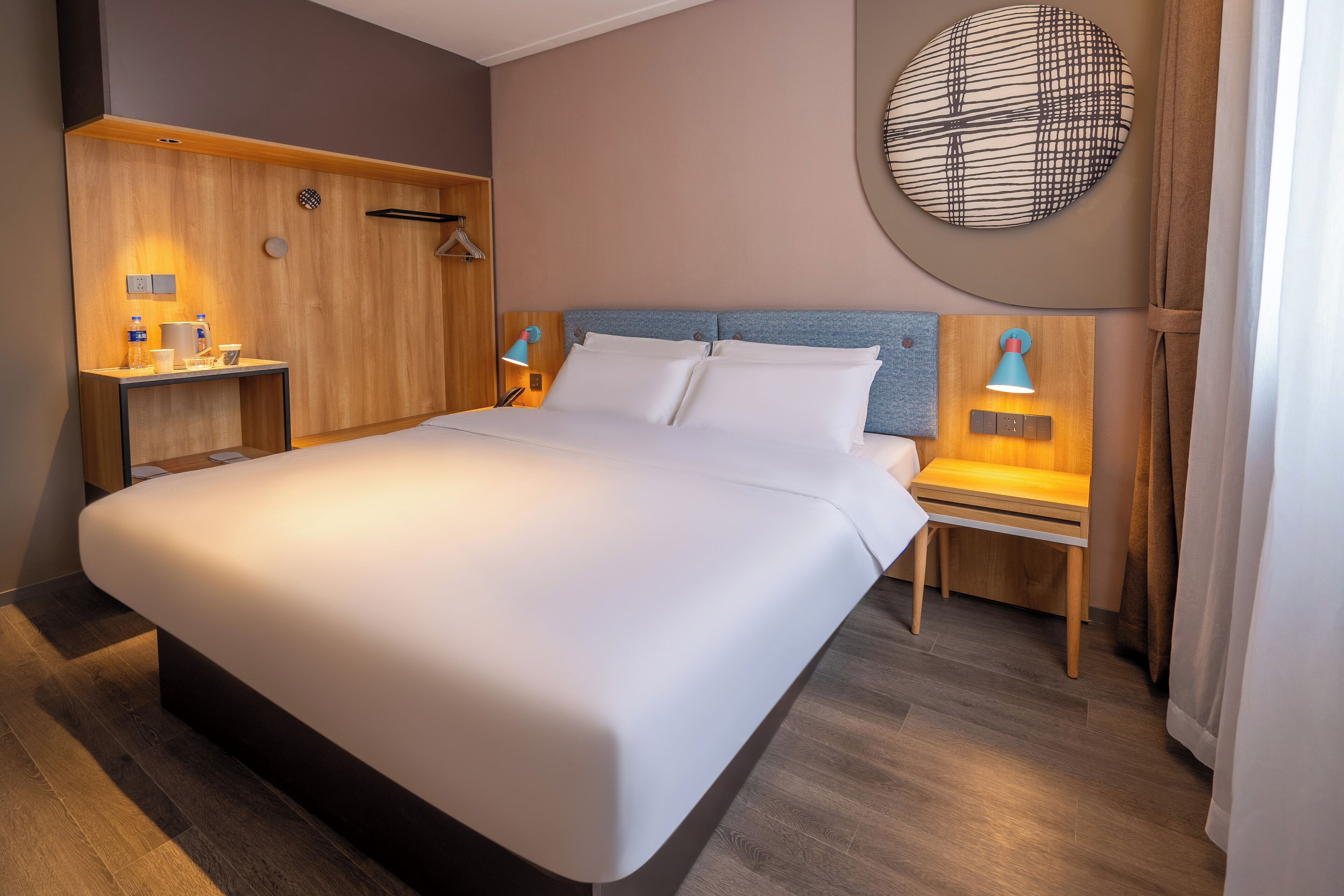 A modern hotel room with a large bed, wooden headboard, bedside tables with lamps, and a round wall decoration.
