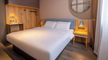 A modern hotel room with a large bed, wooden headboard, bedside tables with lamps, and a round wall decoration.