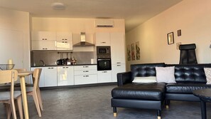 Un salon moderne avec un canapé en cuir noir, un coin repas avec une table et des chaises en bois, et une cuisine avec des meubles et des appareils électroménagers blancs.