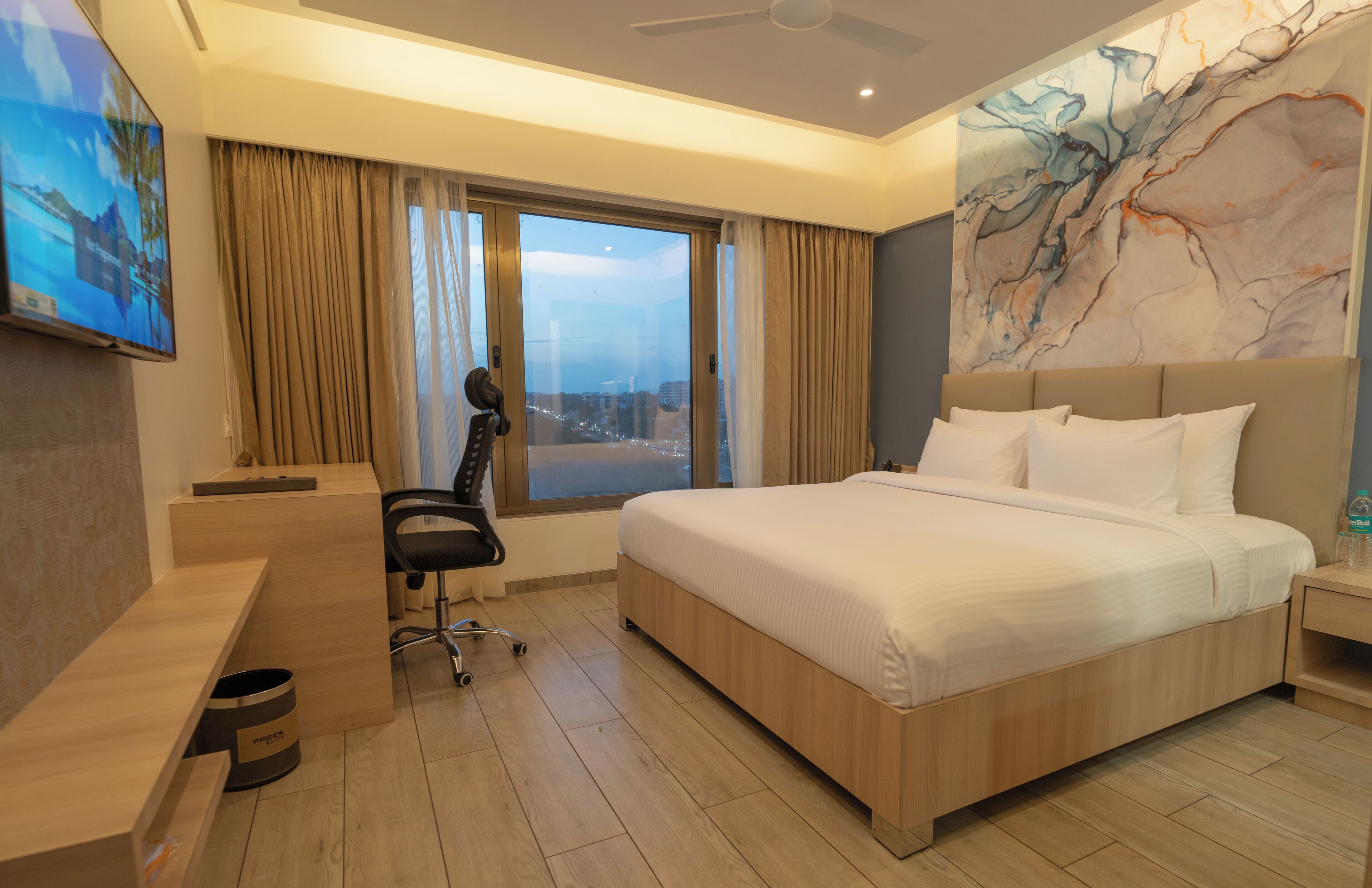 A hotel room with a bed, a desk, a chair, a large window with curtains, and a city view.