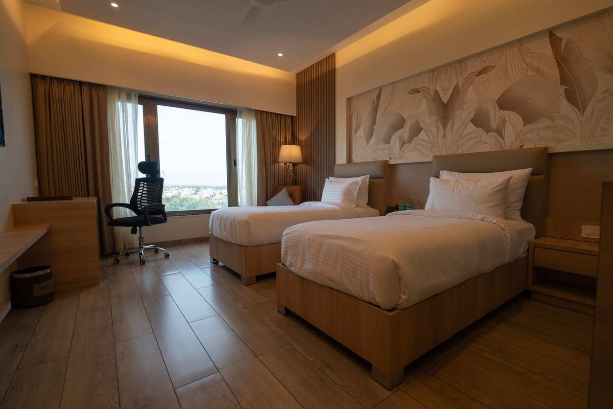 Habitación de hotel con dos camas, un escritorio, una silla y un amplio ventanal con cortinas.