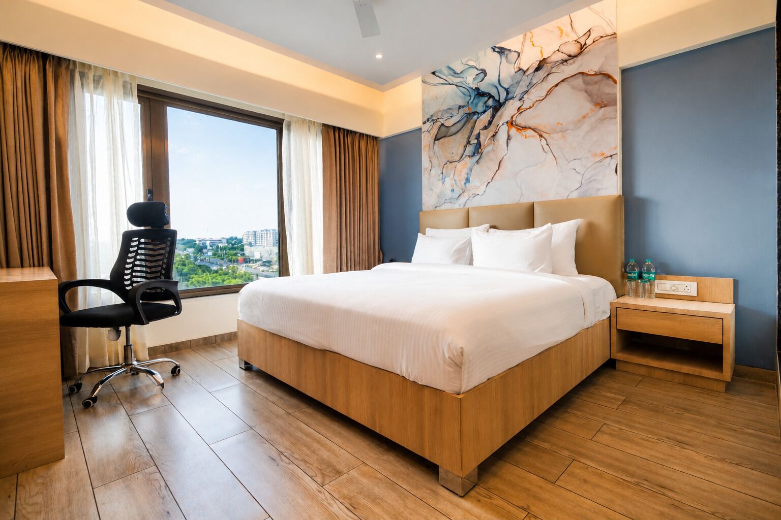 A hotel room with a large bed, a desk, a chair, a nightstand, and a view of the city.