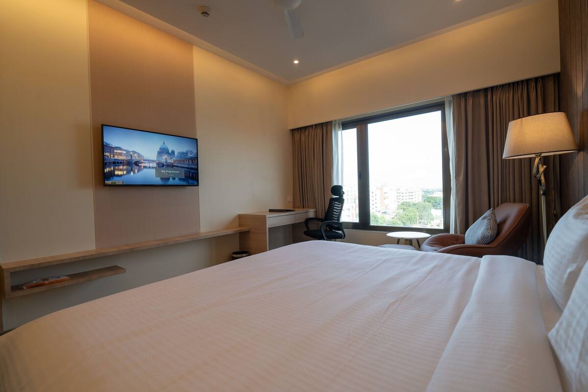 Habitación de hotel con una cama grande, un escritorio, una silla, un televisor montado en la pared y una ventana con cortinas.