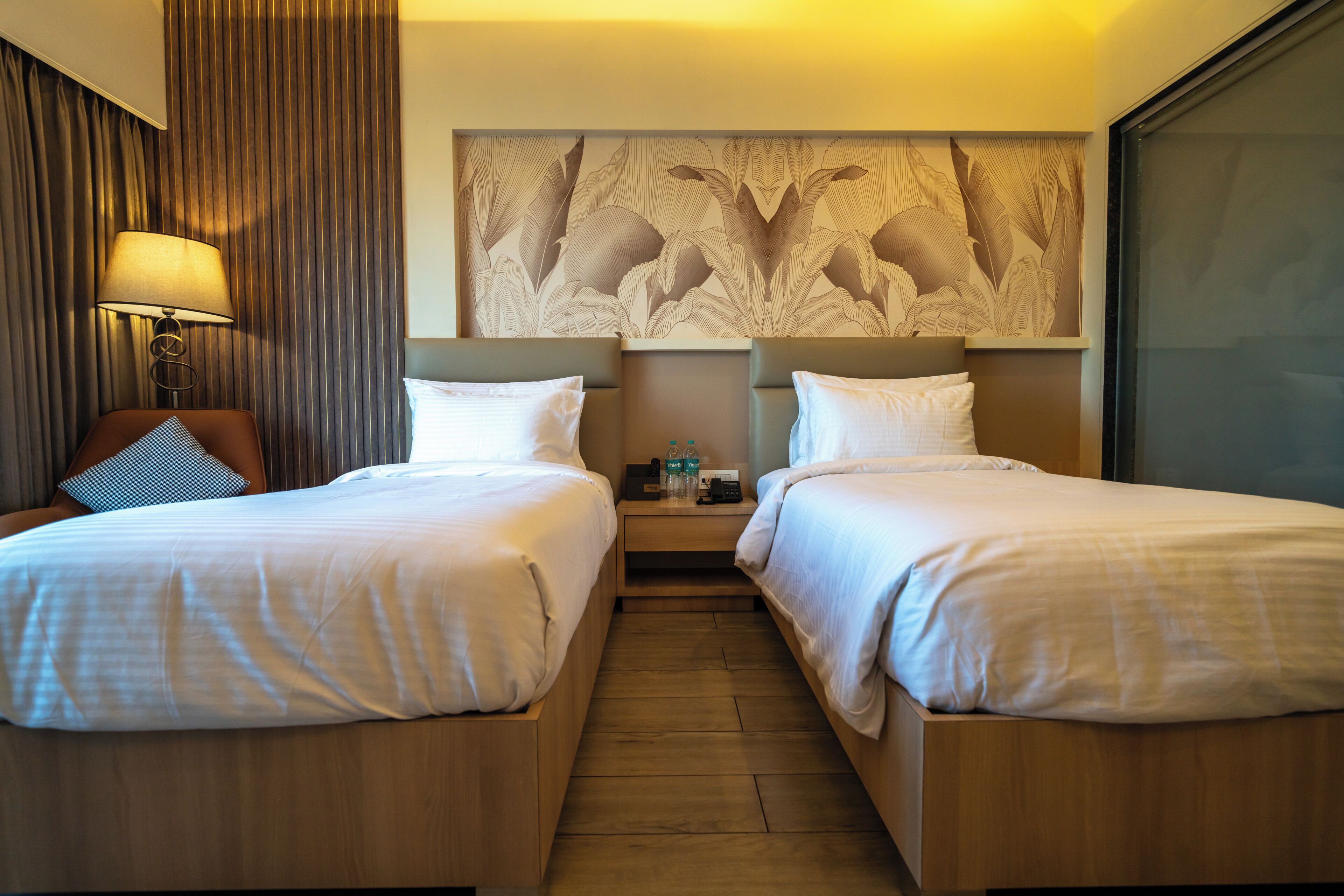Quarto de hotel com duas camas, uma mesa de cabeceira, uma luminária e uma obra de arte na parede.