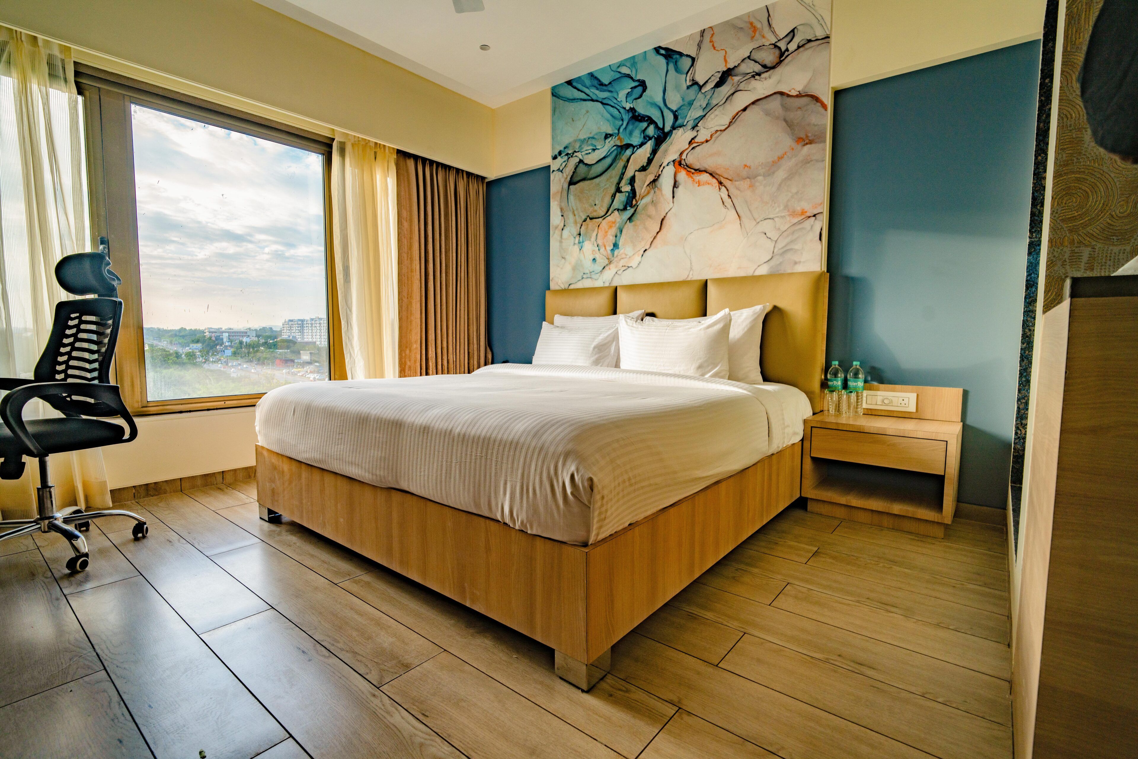 Habitación de hotel con una cama grande, un escritorio, una silla, una mesita de noche y vistas a la ciudad desde la ventana.
