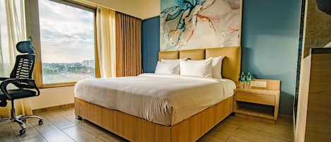 Habitación de hotel con una cama grande, un escritorio, una silla, una mesita de noche y vistas a la ciudad desde la ventana.