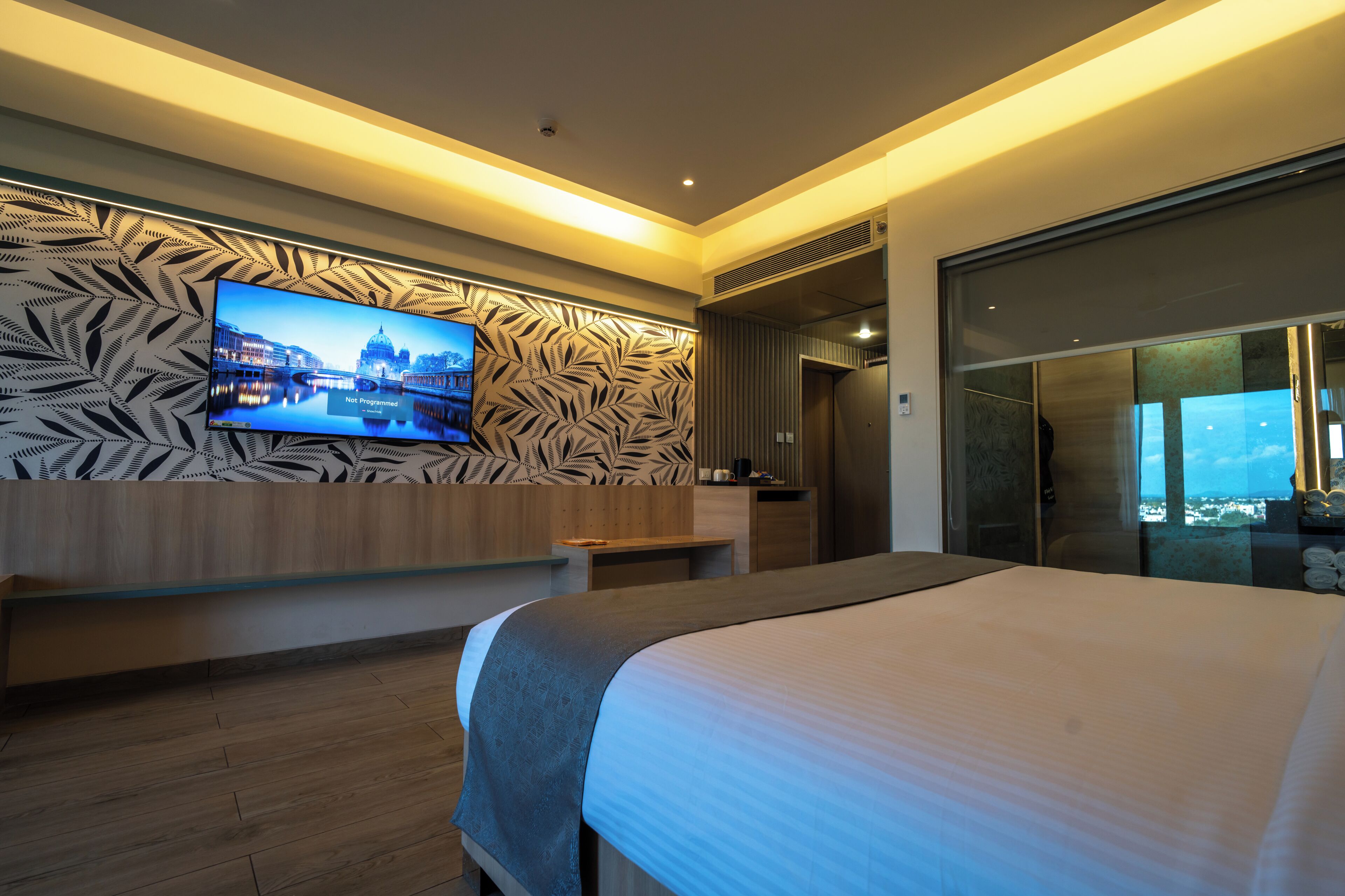 A modern hotel room with a large bed, a television mounted on the wall, and a view of the cityscape.