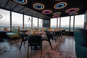 Un comedor moderno con mesas redondas, sillas y vistas panorámicas de la ciudad a través de amplios ventanales.