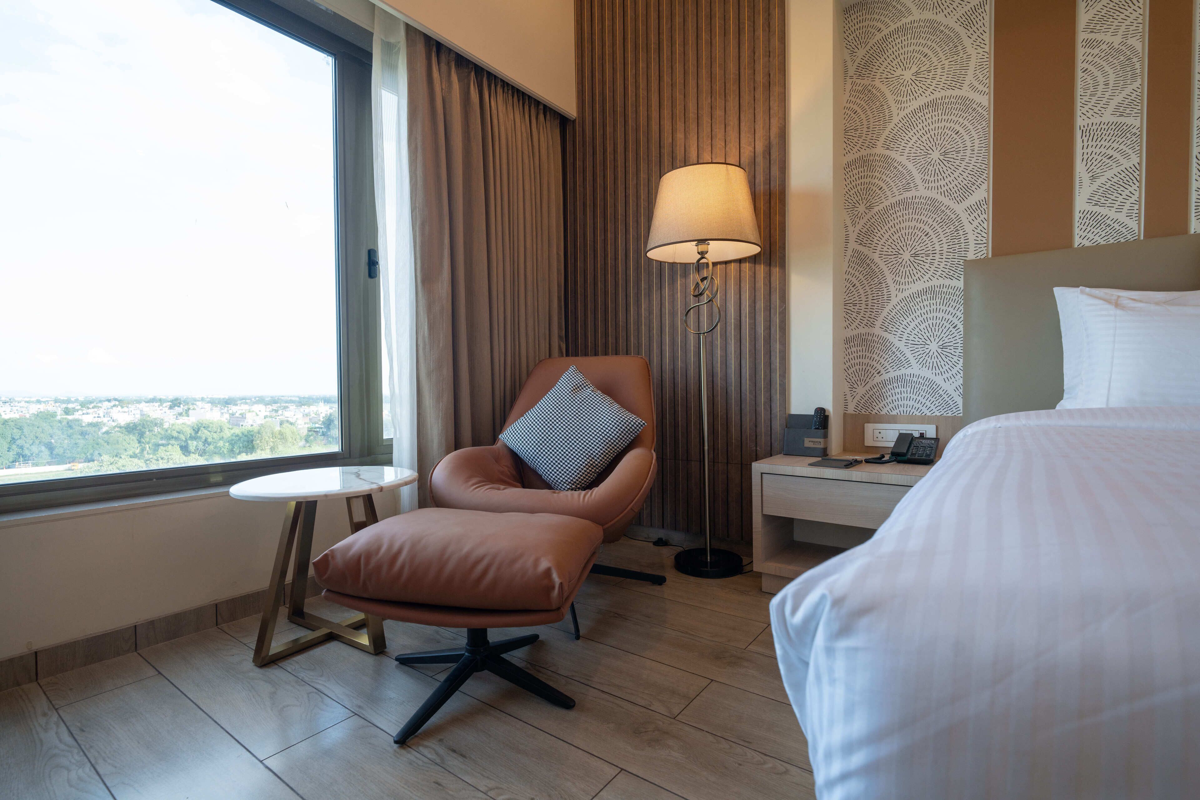 A hotel room with a bed, a chair with a pillow, a small table, a lamp, and a view of the city through a window.