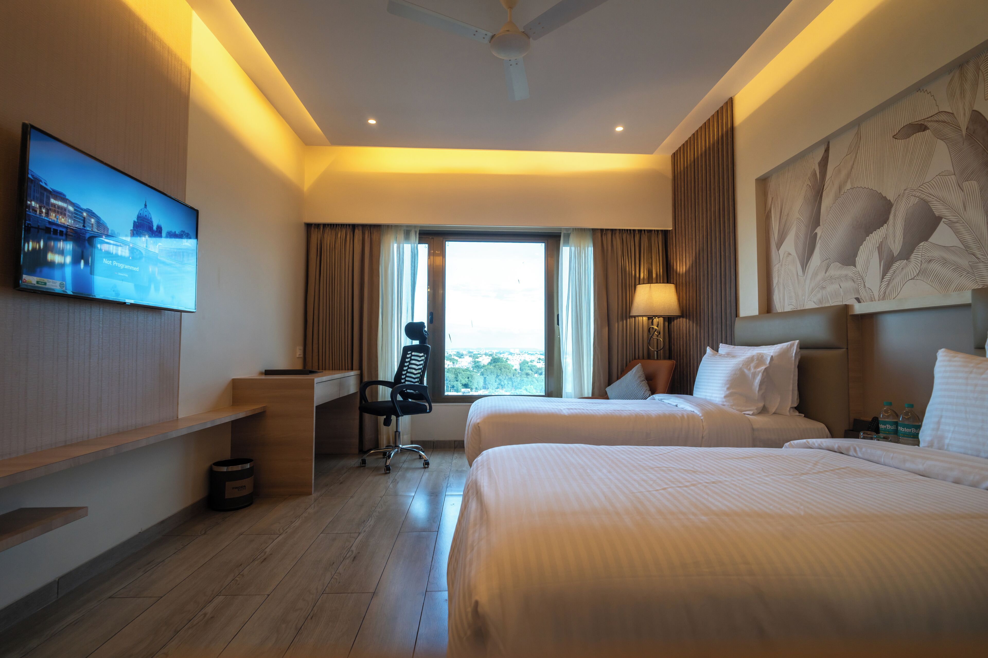 Quarto de hotel com duas camas, uma mesa, uma televisão e uma janela com cortinas.