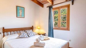 A neatly made bed with a white quilt, pillows, and a folded towel. There is a small table with a lamp and a tray of items. A window with curtains is visible, and a framed picture hangs on the wall. - Villa Rosa (Pollença)