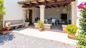 A covered outdoor seating area with stone benches and potted plants, leading to a patio with a table and chairs. - Villa Rosa (Pollença)