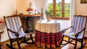A room with two rocking chairs, a table with a tablecloth, and a window with a view of greenery. - Villa Rosa (Pollença)