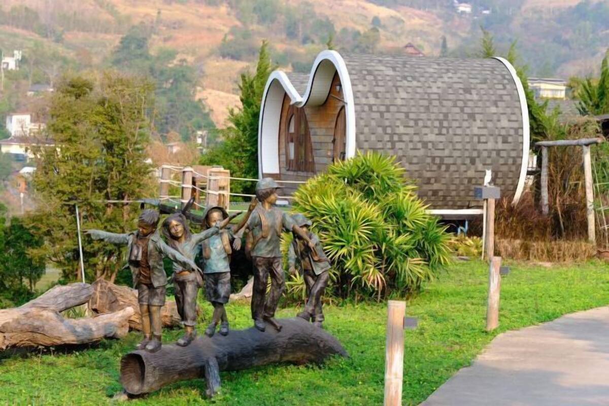 Um edifício cilíndrico peculiar com telhado inclinado, rodeado por uma área ajardinada com uma escultura de crianças e uma grande árvore.