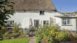 Une petite maison blanche au toit de chaume, un petit jardin avec des plantes en pot et une table avec des chaises à l’extérieur.
