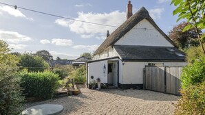 Une maison blanche traditionnelle, avec un toit de chaume, une allée en gravier et un jardin agrémenté de plantes en pot et d’une sculpture.