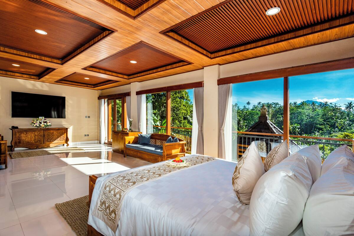 A spacious bedroom with a large bed, a sofa, a TV, and a balcony with a view of the forest.