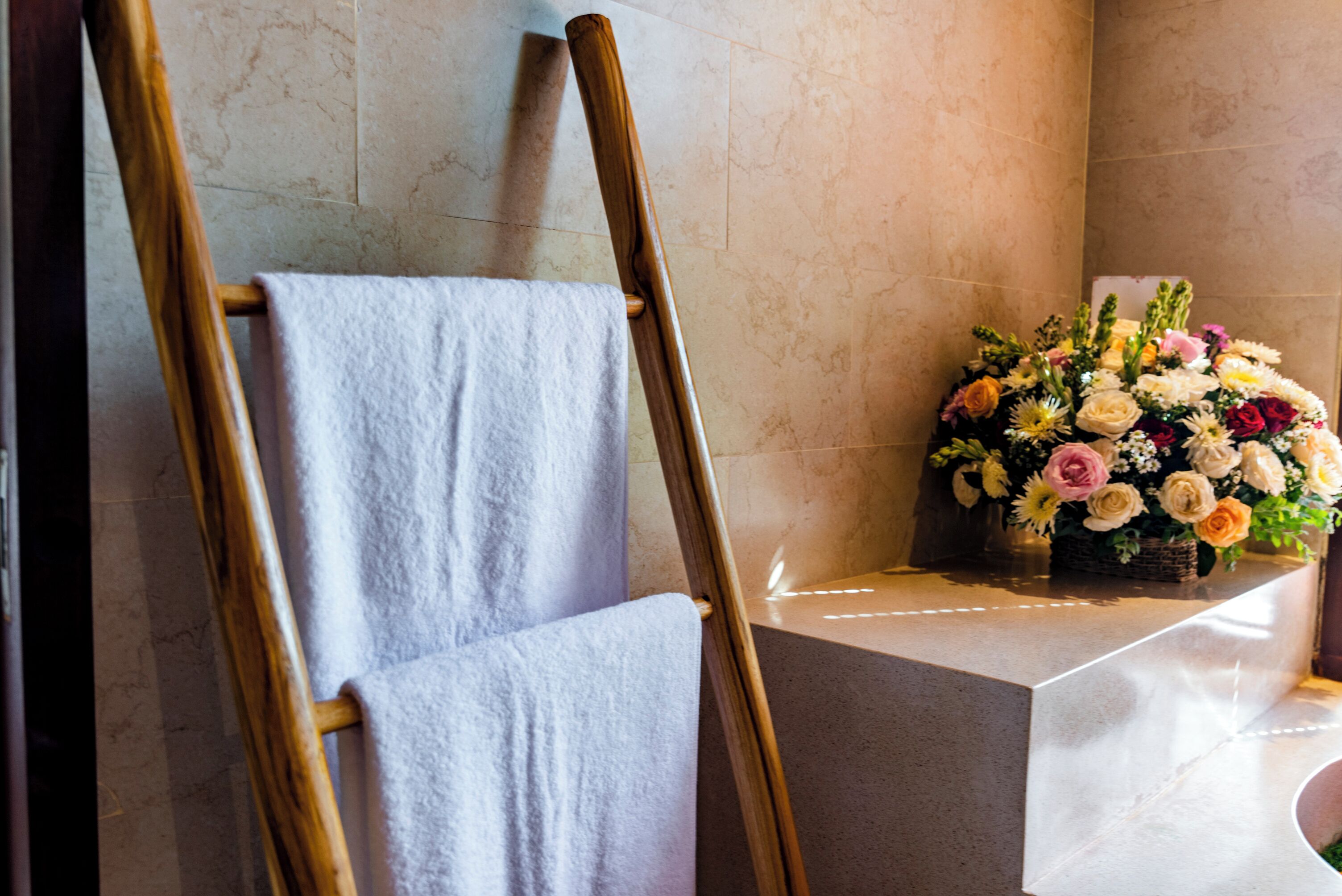 A wooden easel with a white towel draped over it, a floral arrangement on a pedestal, and a tiled wall in the background.
