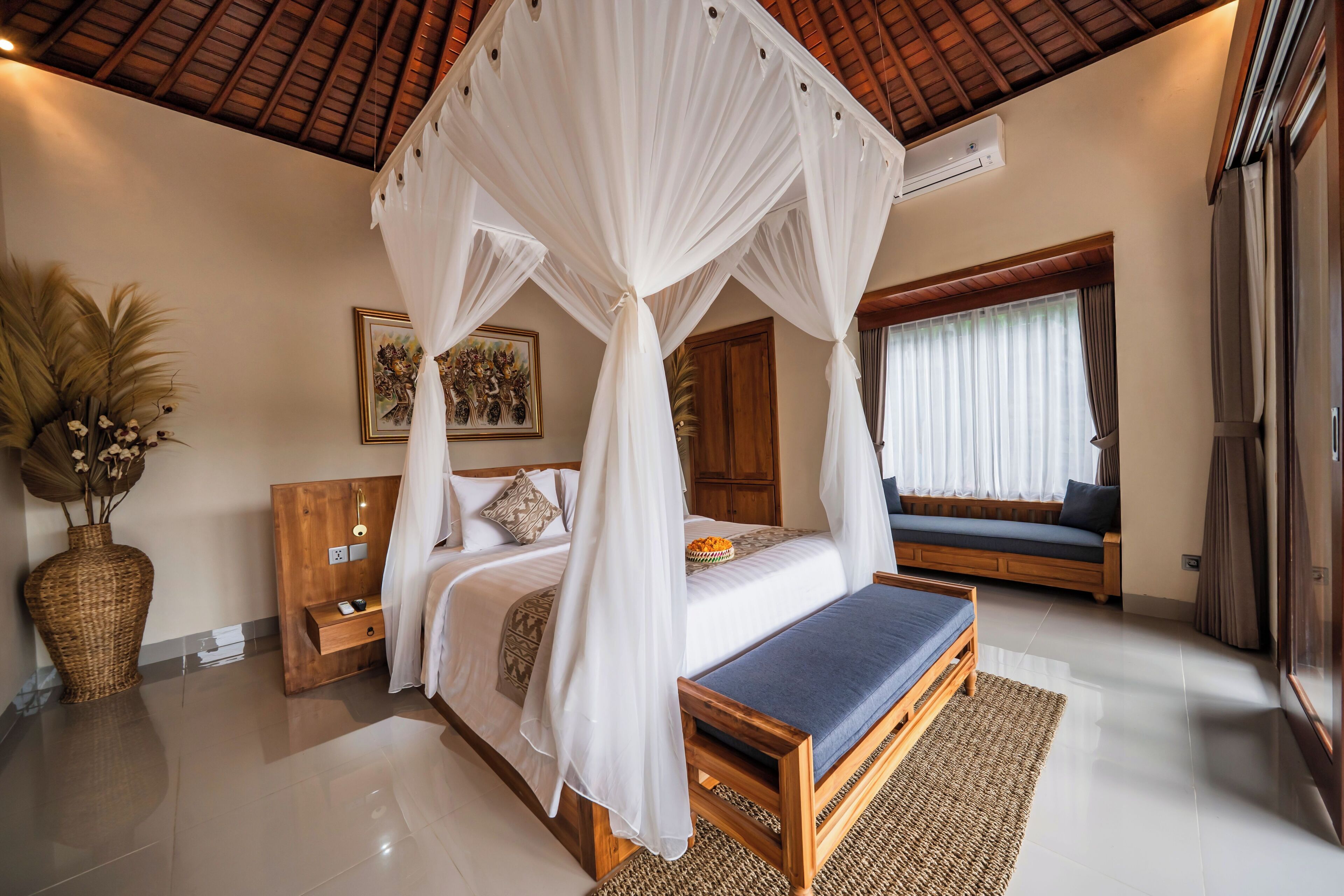 A bedroom with a canopy bed, a wooden bench, a bedside table, a large vase with dried flowers, and a window with curtains.