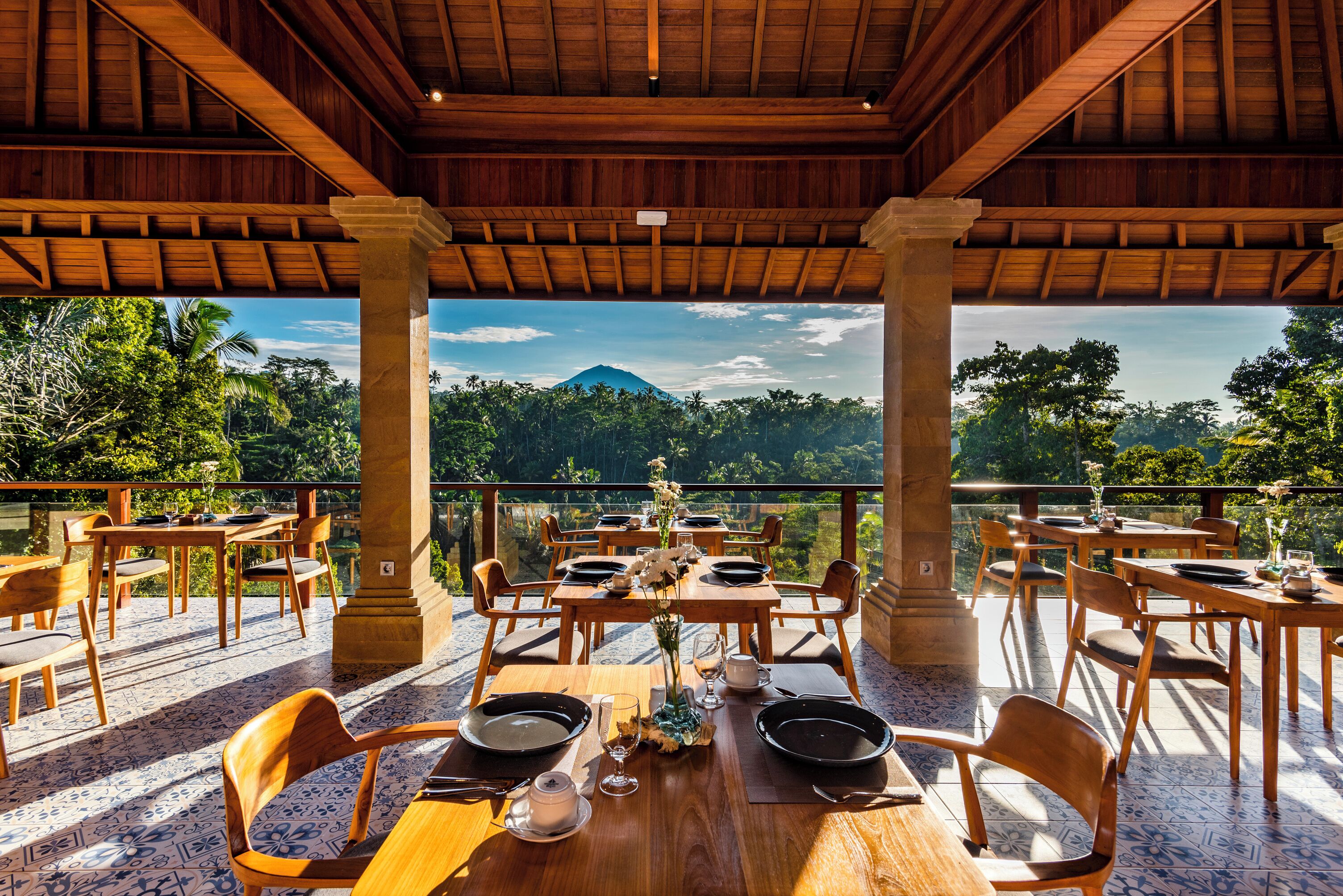 An outdoor dining area with wooden tables and chairs, a view of a mountain, and lush greenery.