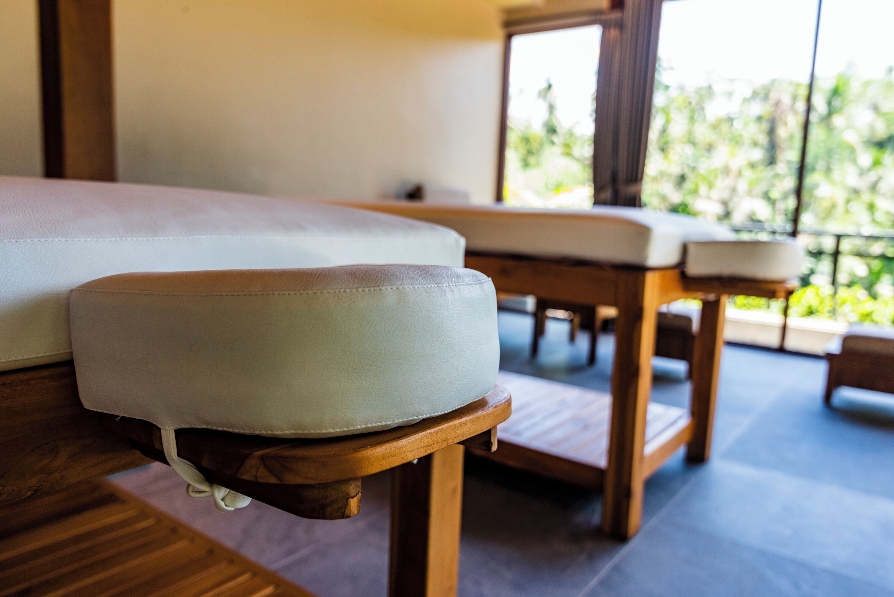 Una mesa de masaje con cojín, un banco de madera y una ventana con vistas a follaje.