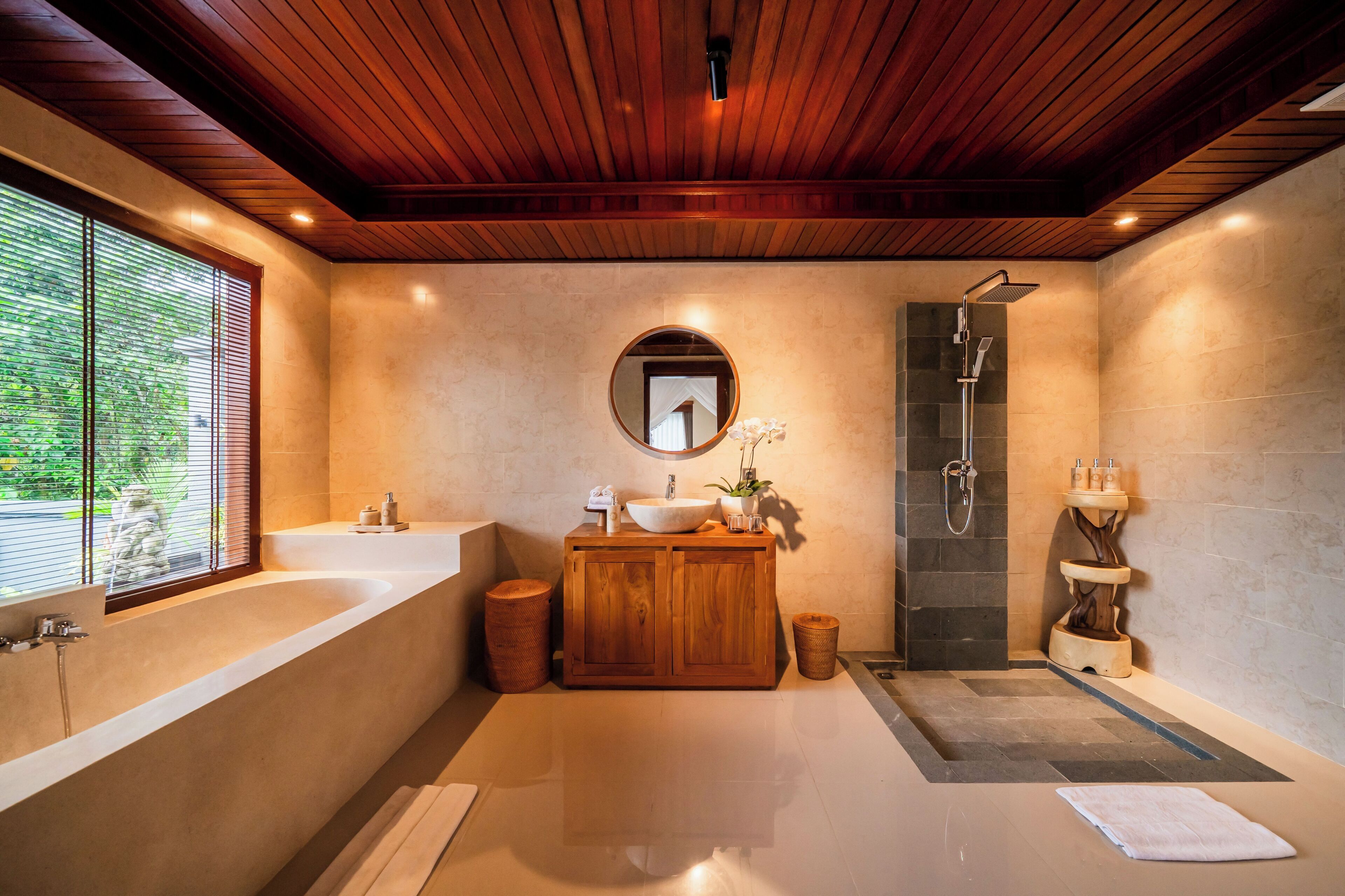 A modern bathroom with a wooden ceiling, a large bathtub, a round mirror, a sink with a wooden vanity, and a shower area.
