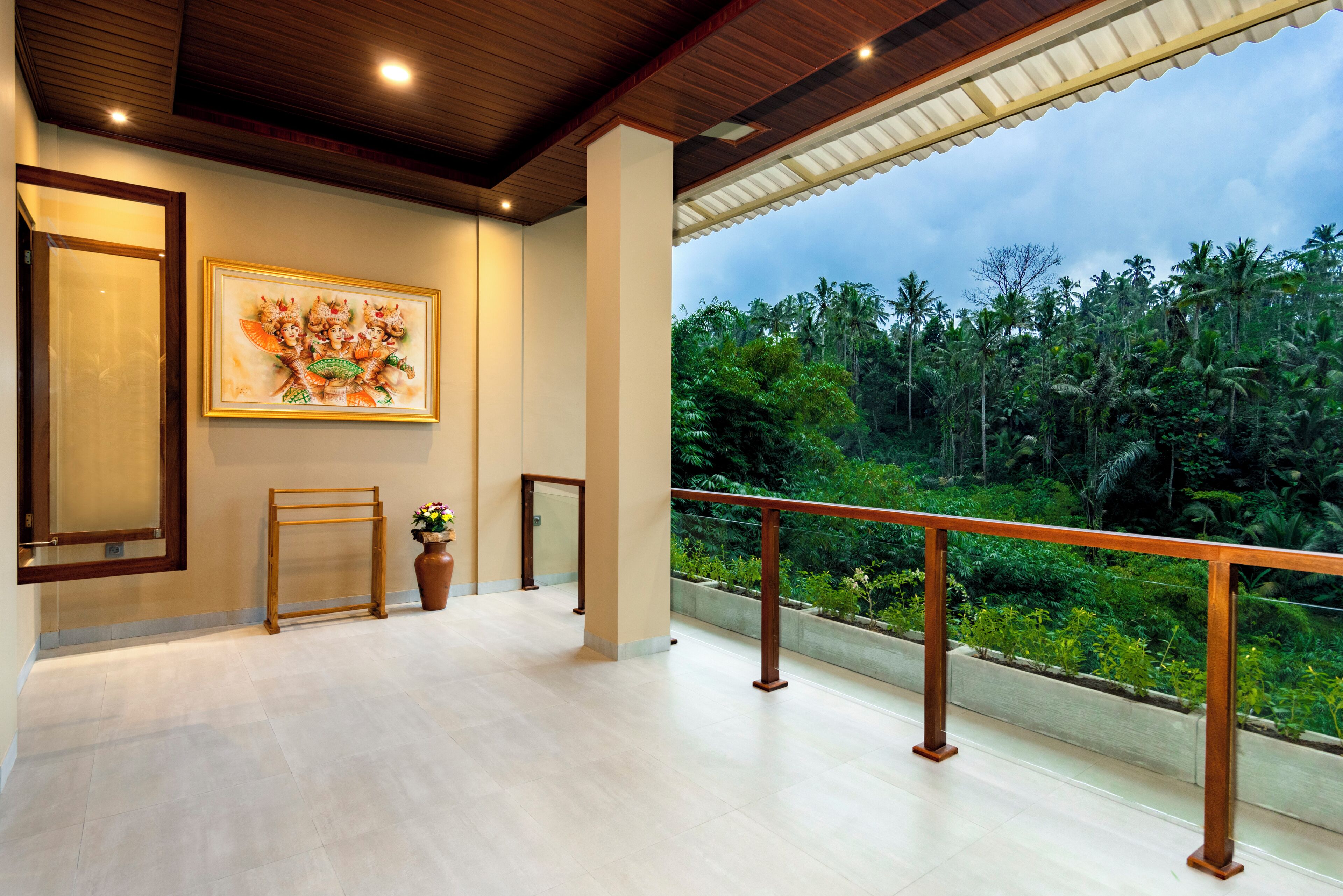 A balcony with a view of a tropical landscape, a framed artwork on the wall, and a wooden rack.