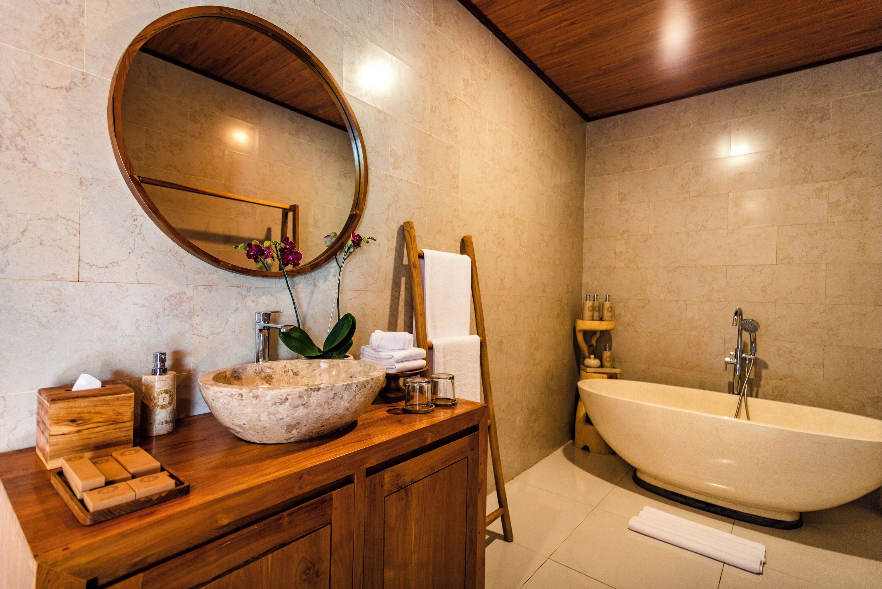 A bathroom with a wooden vanity, a large oval mirror, a freestanding bathtub, and a marble sink.
