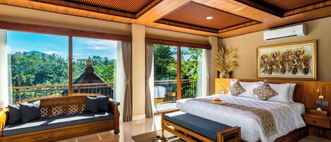 A spacious bedroom with a large bed, a wooden bench, and a sofa. It features a balcony with a view of lush greenery and a traditional structure.
