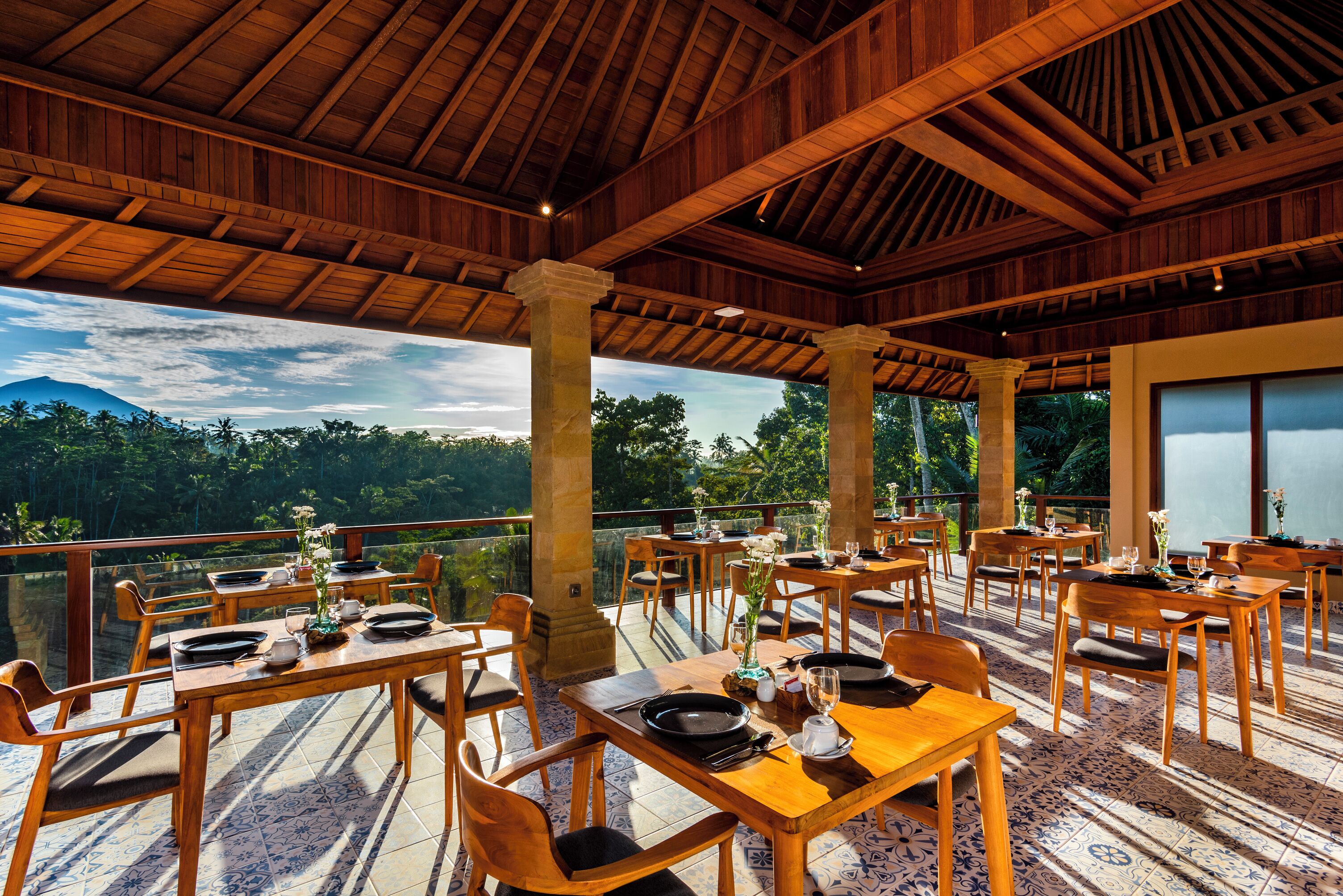 Un espacio de comedor al aire libre con mesas y sillas de madera, con un fondo de vegetación exuberante y una montaña.