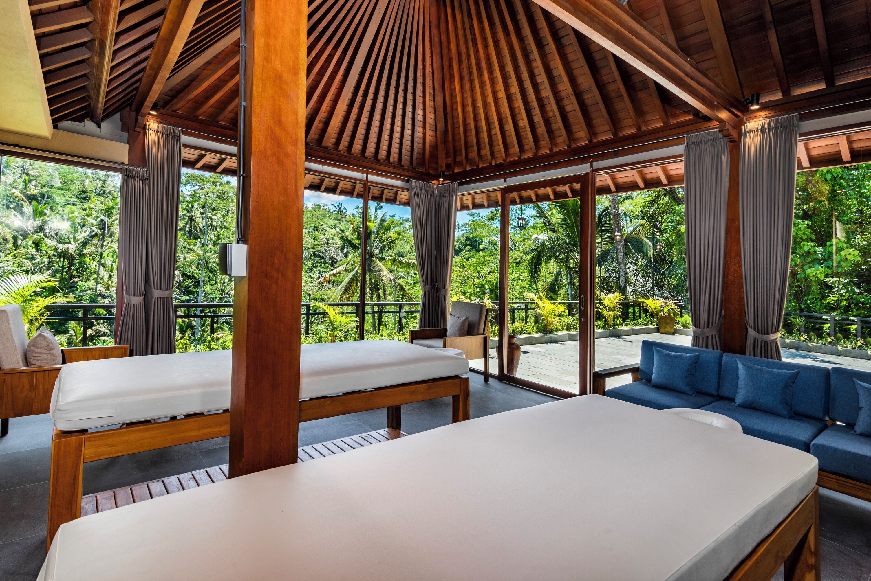 A spacious room with a wooden ceiling and large windows offering a view of lush greenery. There are two beds and a blue sofa.
