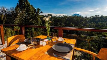A dining table set with plates, cutlery, and a vase of flowers, overlooking a lush landscape with trees and a mountain.
