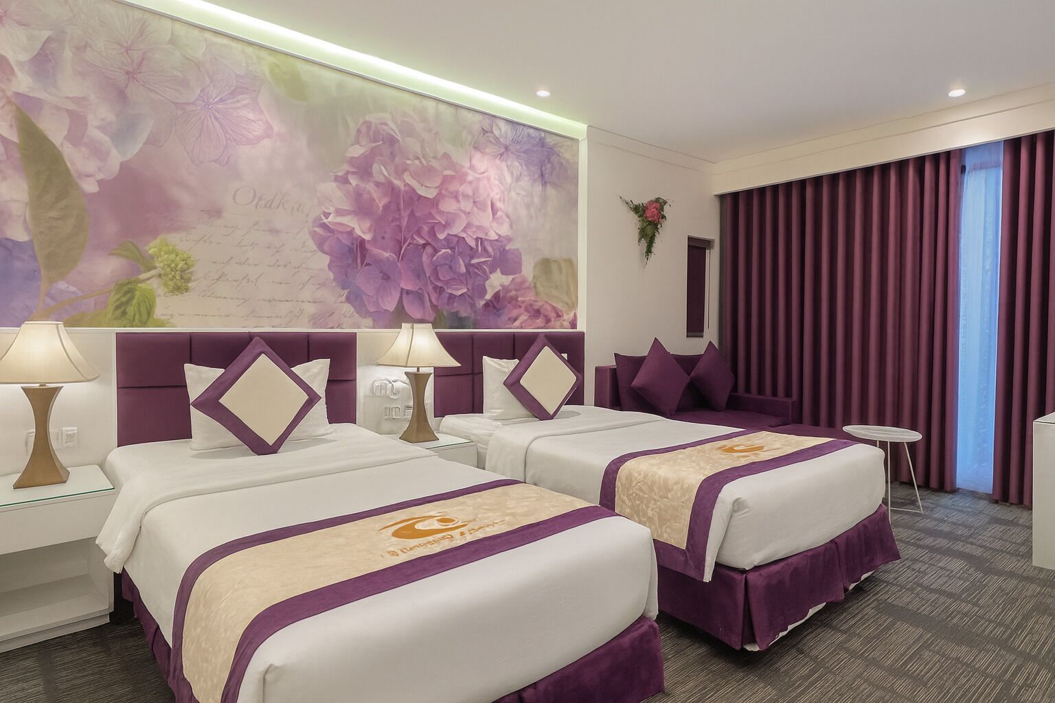 A hotel room with two beds, a large floral mural, a purple headboard, and a view of the city through curtains.