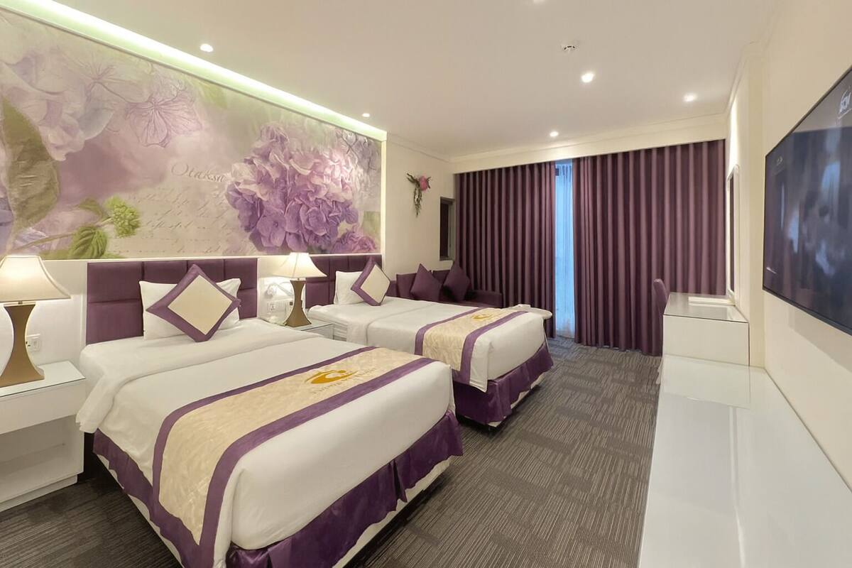 Habitación de hotel con dos camas, un mural floral grande, un televisor de pantalla plana y un ventanal con cortinas moradas.