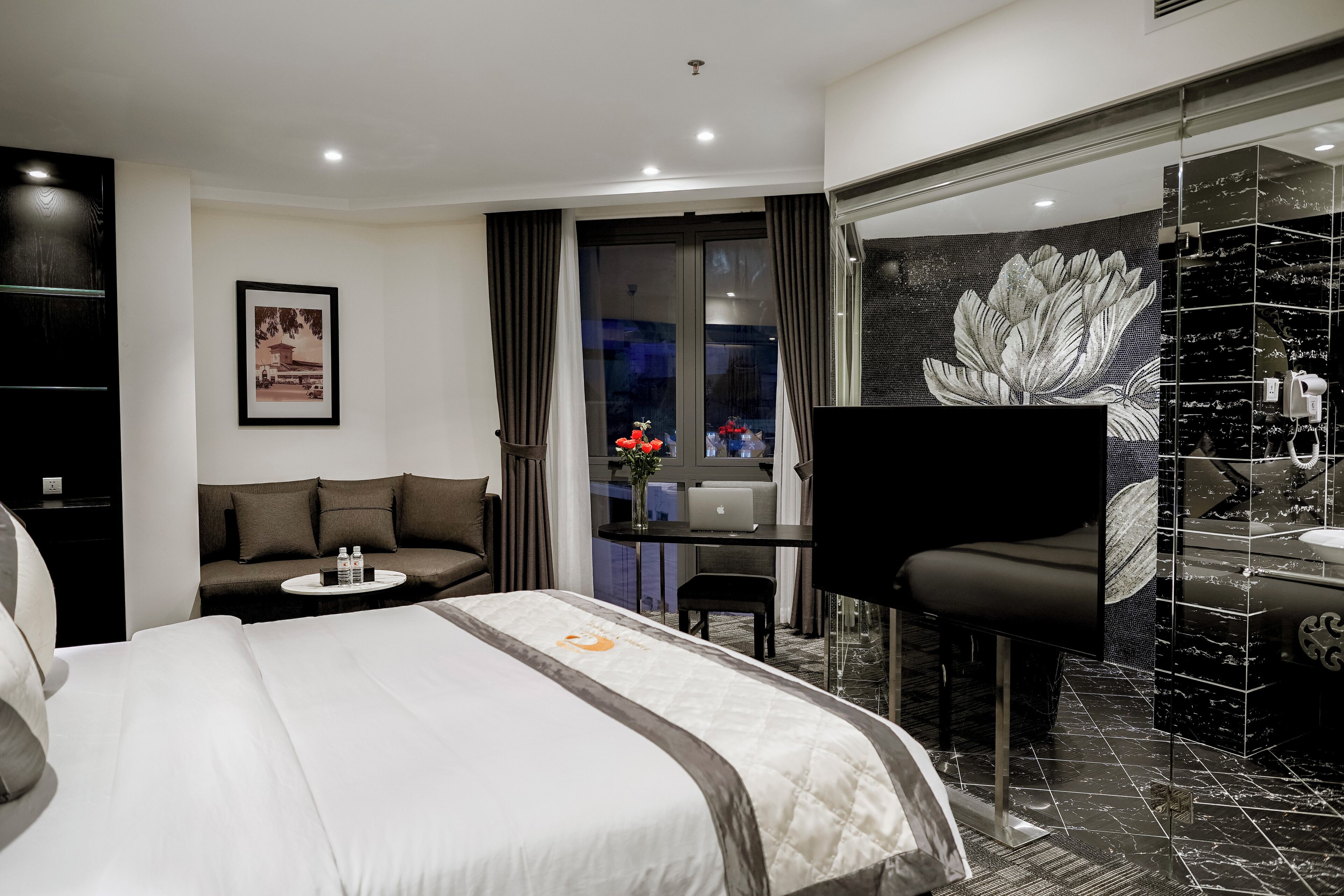 Habitación de hotel moderna con una cama grande, un sofá, un escritorio con una computadora portátil y un baño con un espejo grande y un detalle decorativo en la pared.
