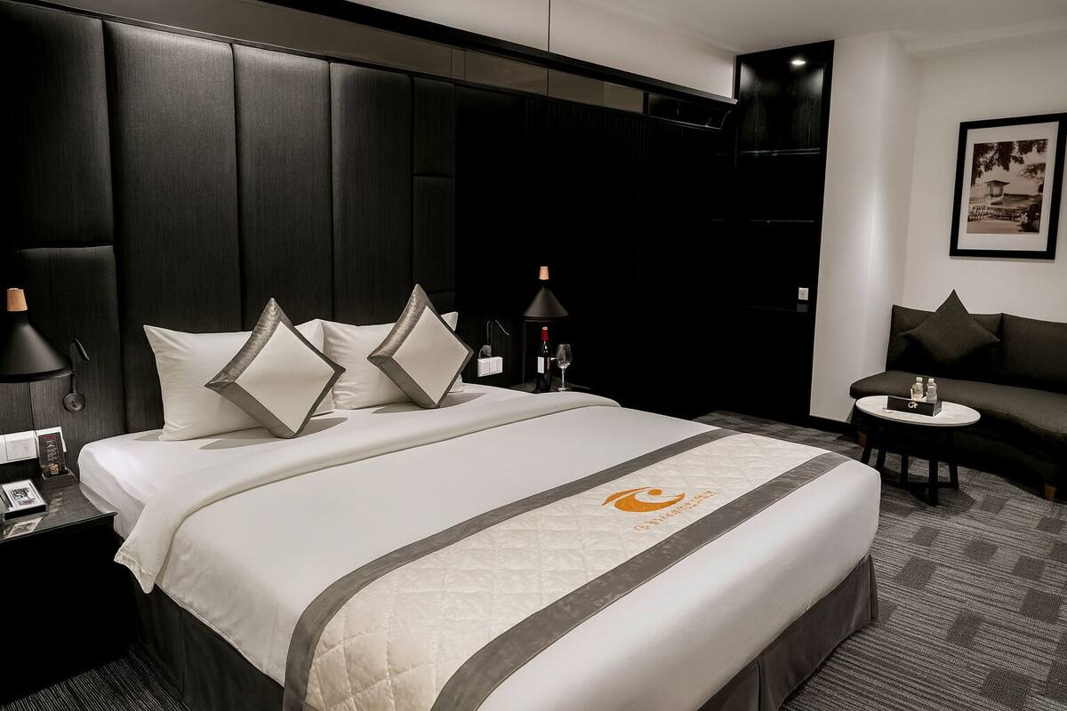Habitación de hotel con una cama grande, cabecera de madera oscura, un pequeño sofá, una mesa redonda y un cuadro en la pared.