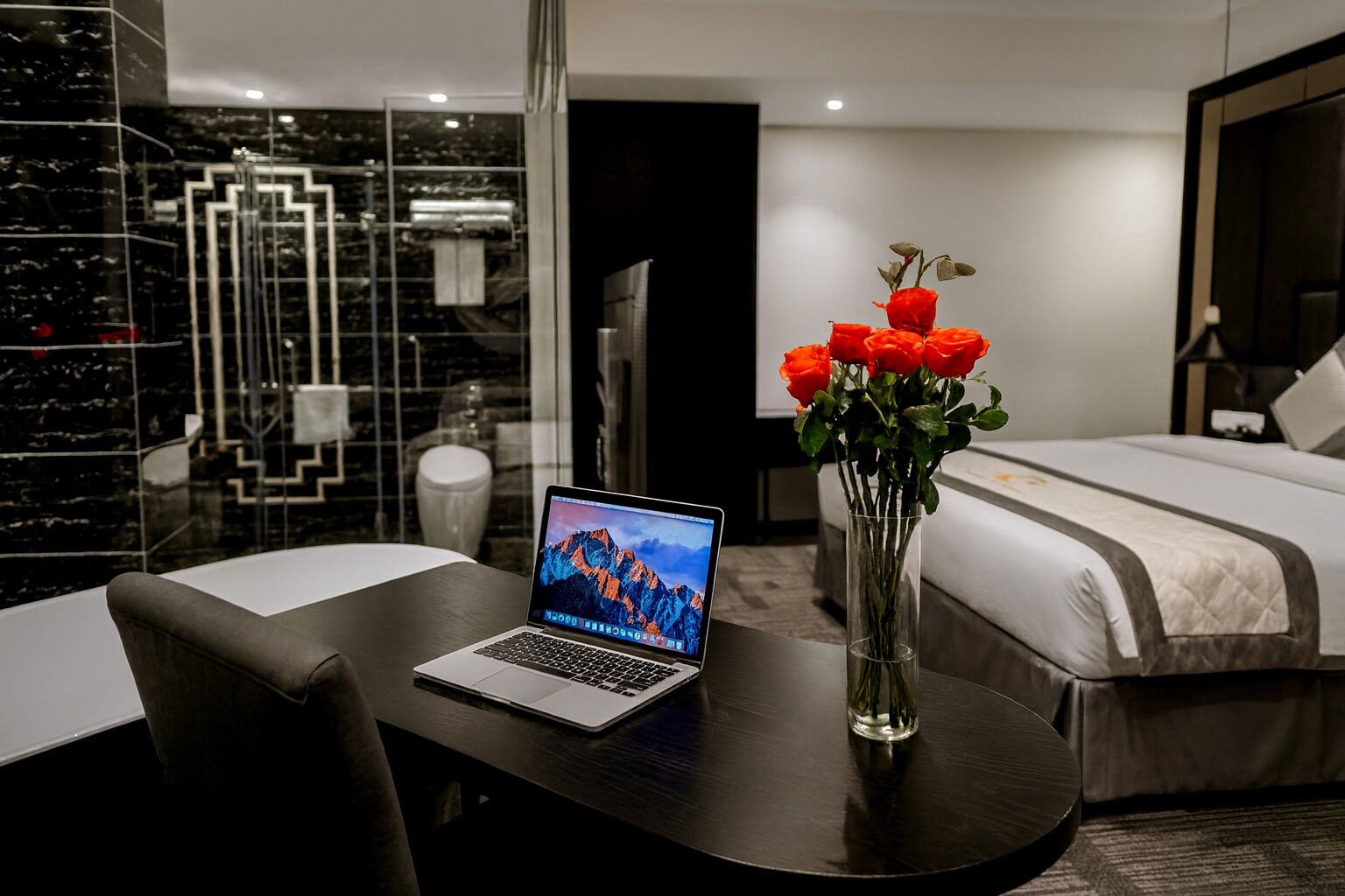A modern hotel room with a bed, a laptop on a desk, and a vase of red roses.