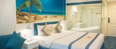 A hotel room with a bed, a nightstand, a desk, a bathroom with a shower, and a large beach mural on the wall.