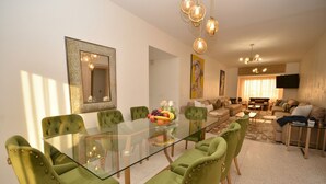 Un comedor con una mesa de vidrio y sillas tapizadas de verde, una sala de estar con un sofá y un televisor, y un cuadro en la pared.