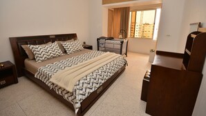 Un dormitorio con cama, mesitas de noche, cómoda, una ventana con cortinas y vistas a edificios.