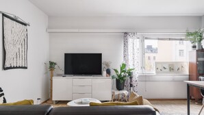 A modern living room with a flat-screen TV, a white cabinet, a dining table, and a window with a view of buildings. - Bright Studio Apt. with Private Balcony  (Helsinki)