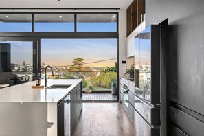 A modern kitchen with a large island, stainless steel appliances, and a view of a cityscape through large windows. - Ocean-View Luxury | Spa, Fire Pit + Ocean Views (Ocean Grove)