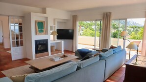 Ein modernes Wohnzimmer mit Sofa, Fernseher, Kamin und großen Fenstern mit Blick auf Grünflächen.