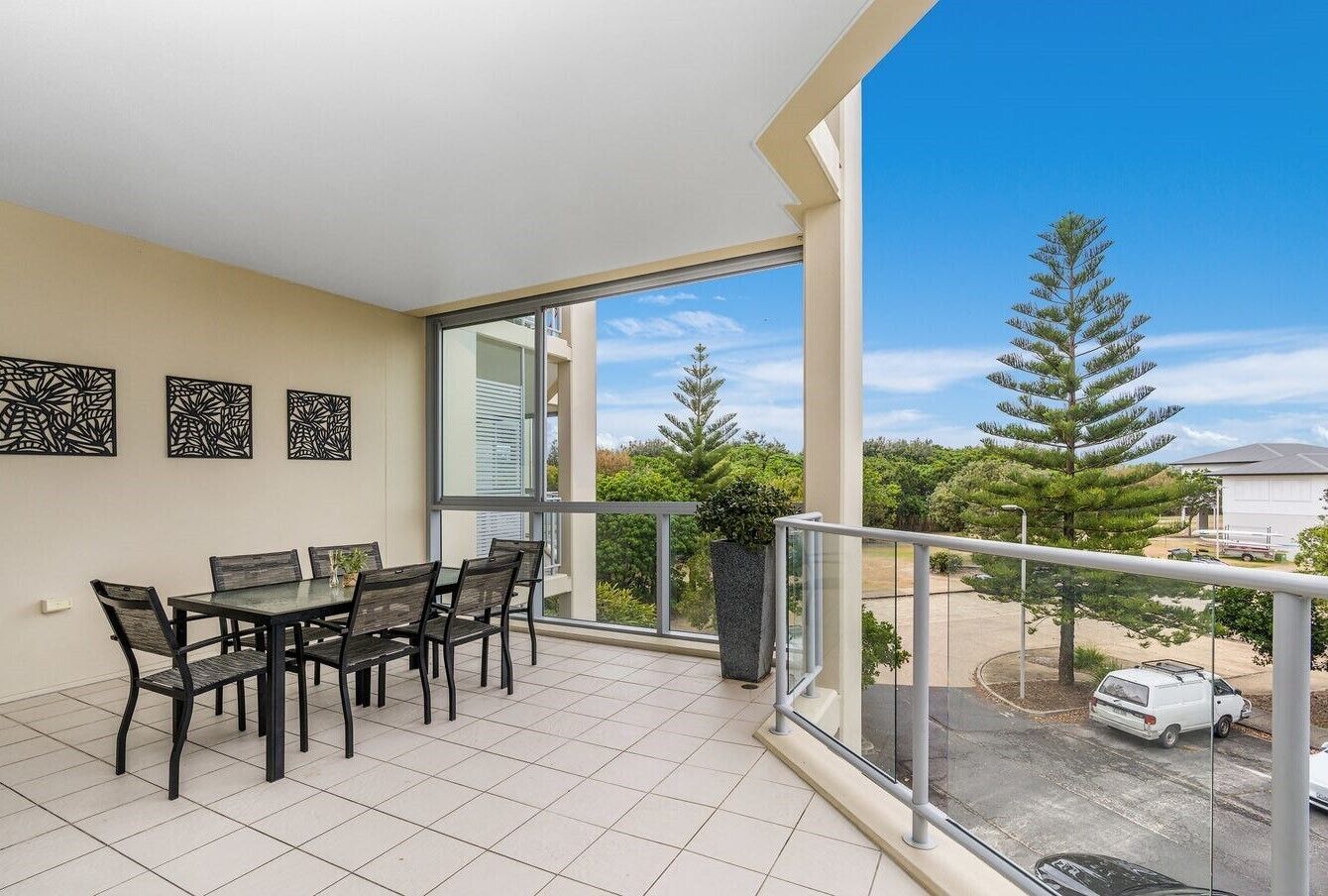 A balcony with a dining table and chairs, a potted plant, and a view of a parking area and trees.
