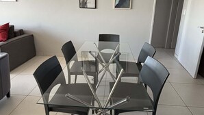 Ein moderner Essbereich mit einem Glastisch und schwarzen Stühlen, ein graues Sofa mit einem roten Kissen sowie zwei gerahmte Kunstwerke an der Wand.