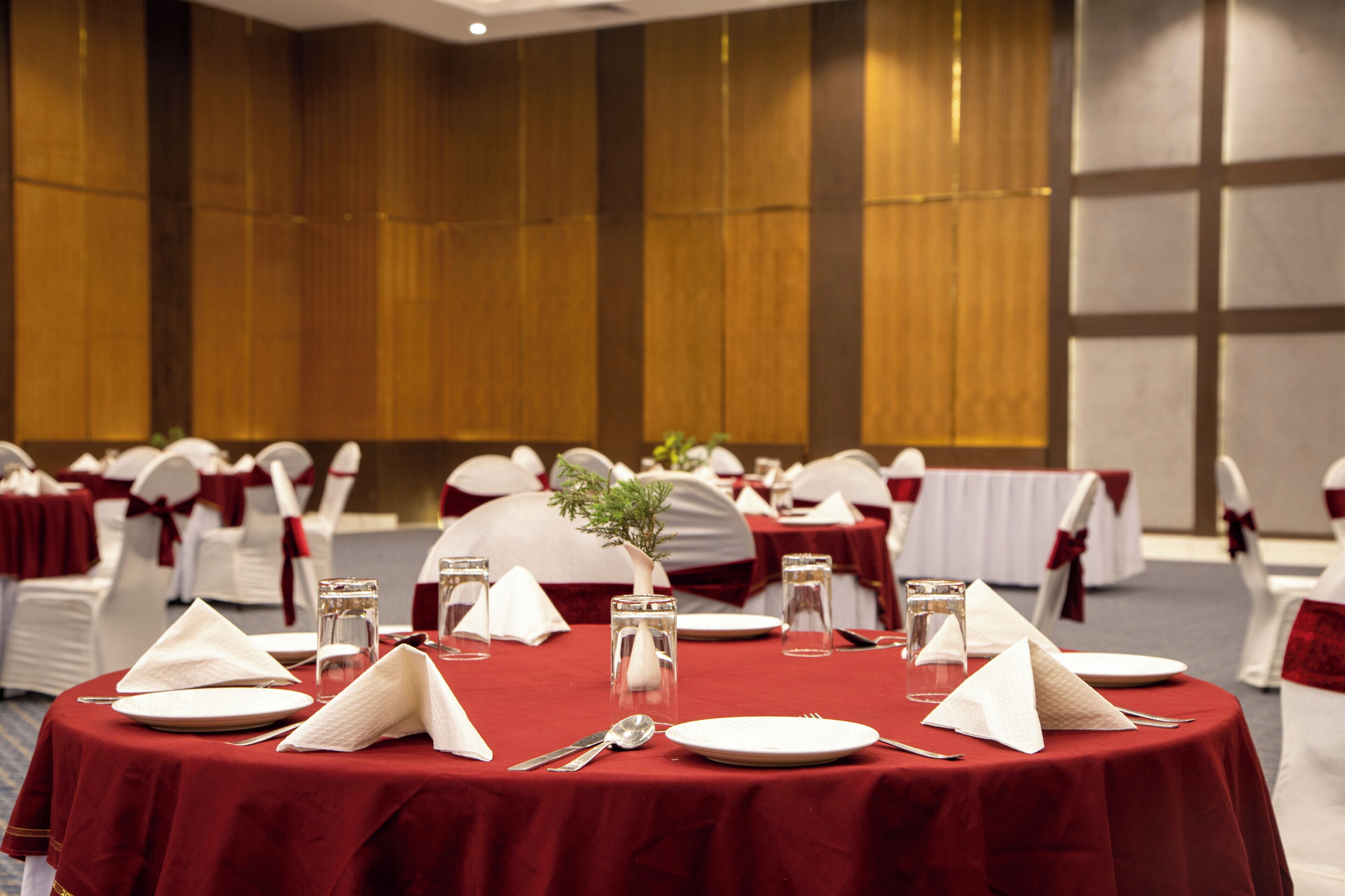 Une salle de banquet avec des tables rondes dressées pour un événement formel, agrémentée de nappes rouges, de serviettes blanches et de verres en cristal.
