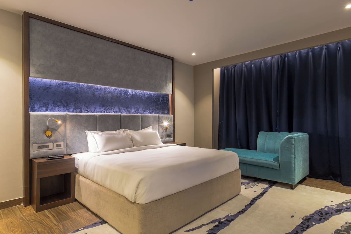 A modern hotel room with a large bed, a headboard with integrated lighting, a teal sofa, and a dark curtain.