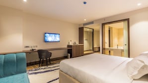 A hotel room with a bed, a desk, a chair, a TV, and a bathroom visible through an open door.